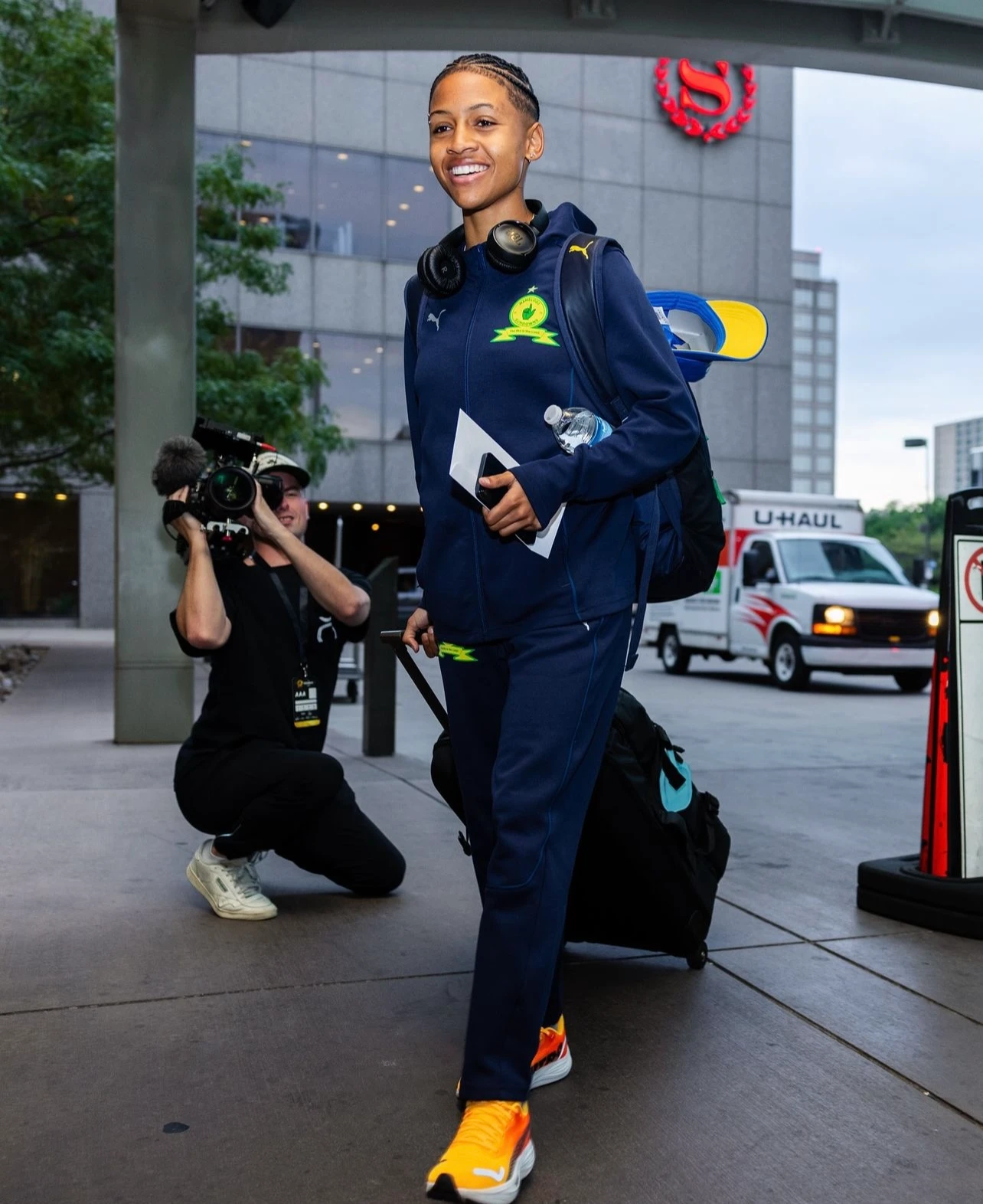 ESCA Alumni Cimone Sauls in blue sports outfit walking with headphones around her neck and pulling a suitcase, while a cameraman films her.