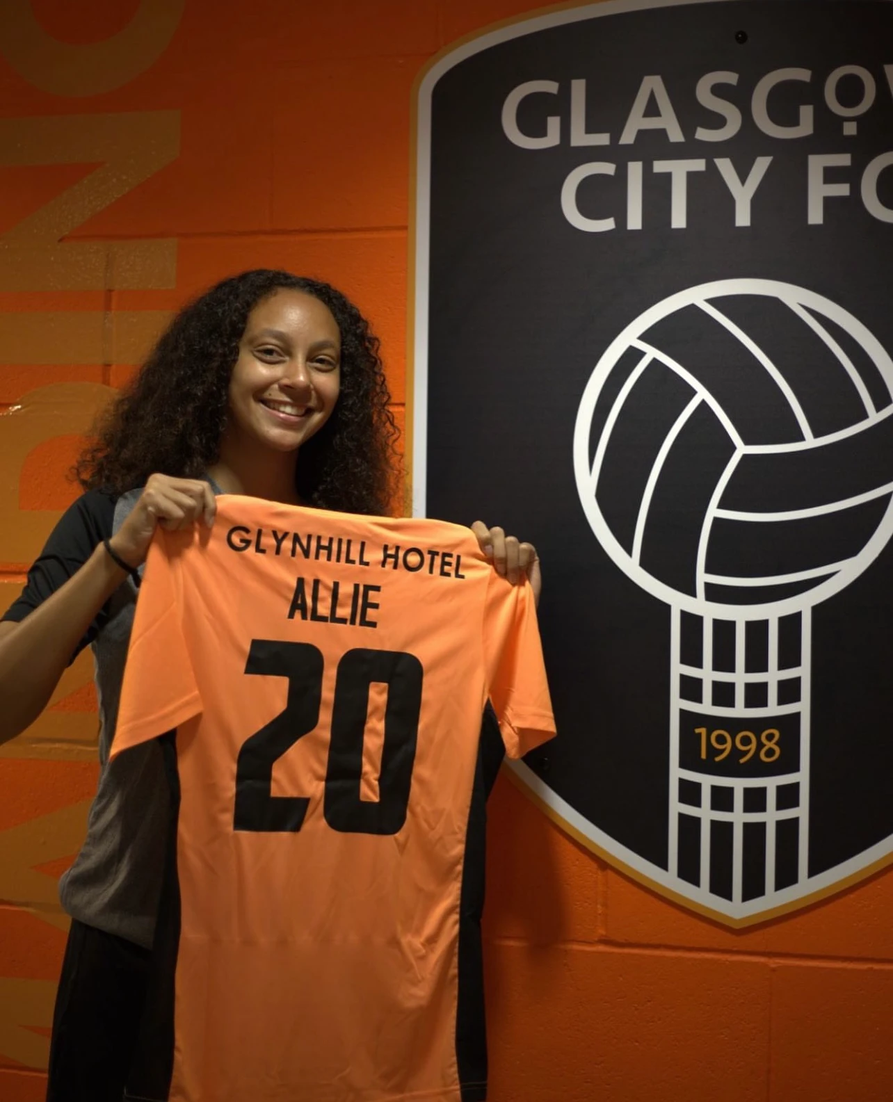 ESCA Alumni Aliyaah Allie holding an orange sports jersey with the name Allie and number 20 in front of a Glasgow City FC logo.