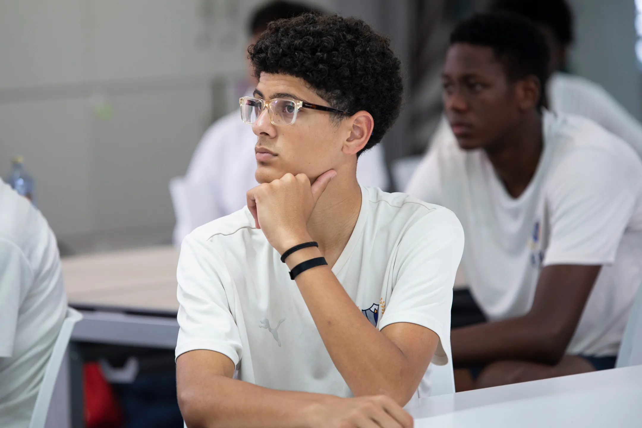ESCA Student with curly hair and glasses sitting at a desk, resting his chin on his hand, attentively looking to the side.