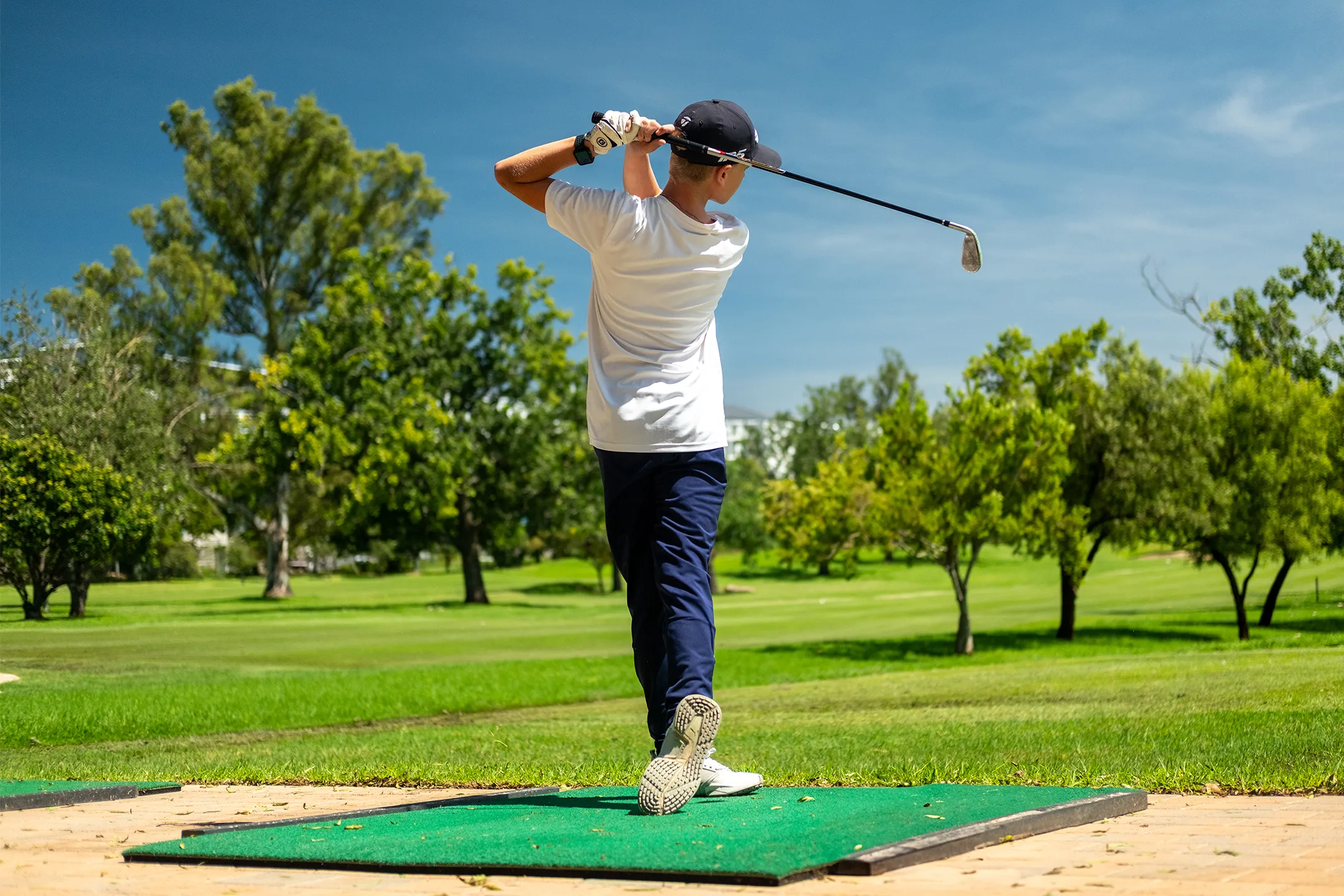 ESCA Student in white shirt and black cap swinging a golf club on a green outdoor golf range.