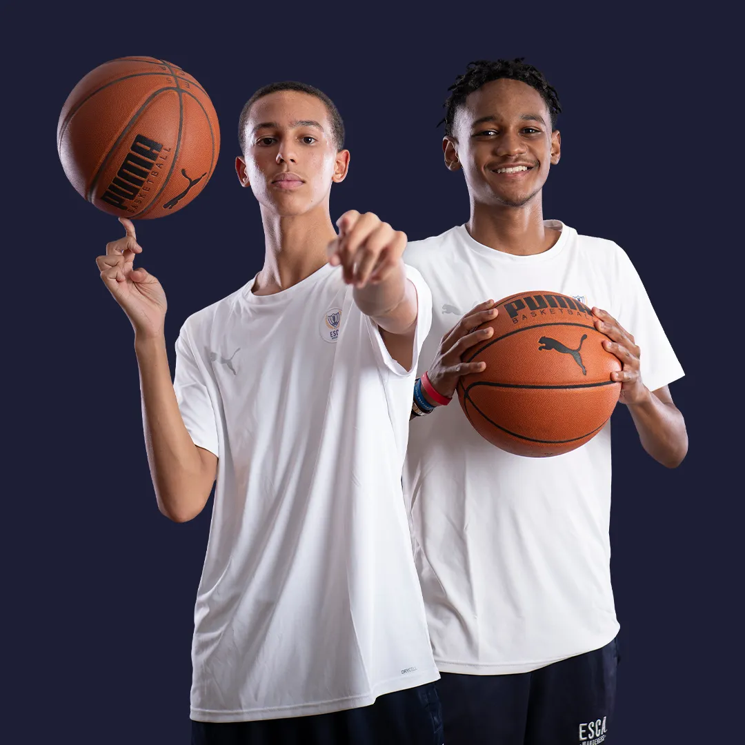 Two young men in white shirts holding basketballs against a dark background, one spinning a ball on his finger and the other smiling while holding a basketball.