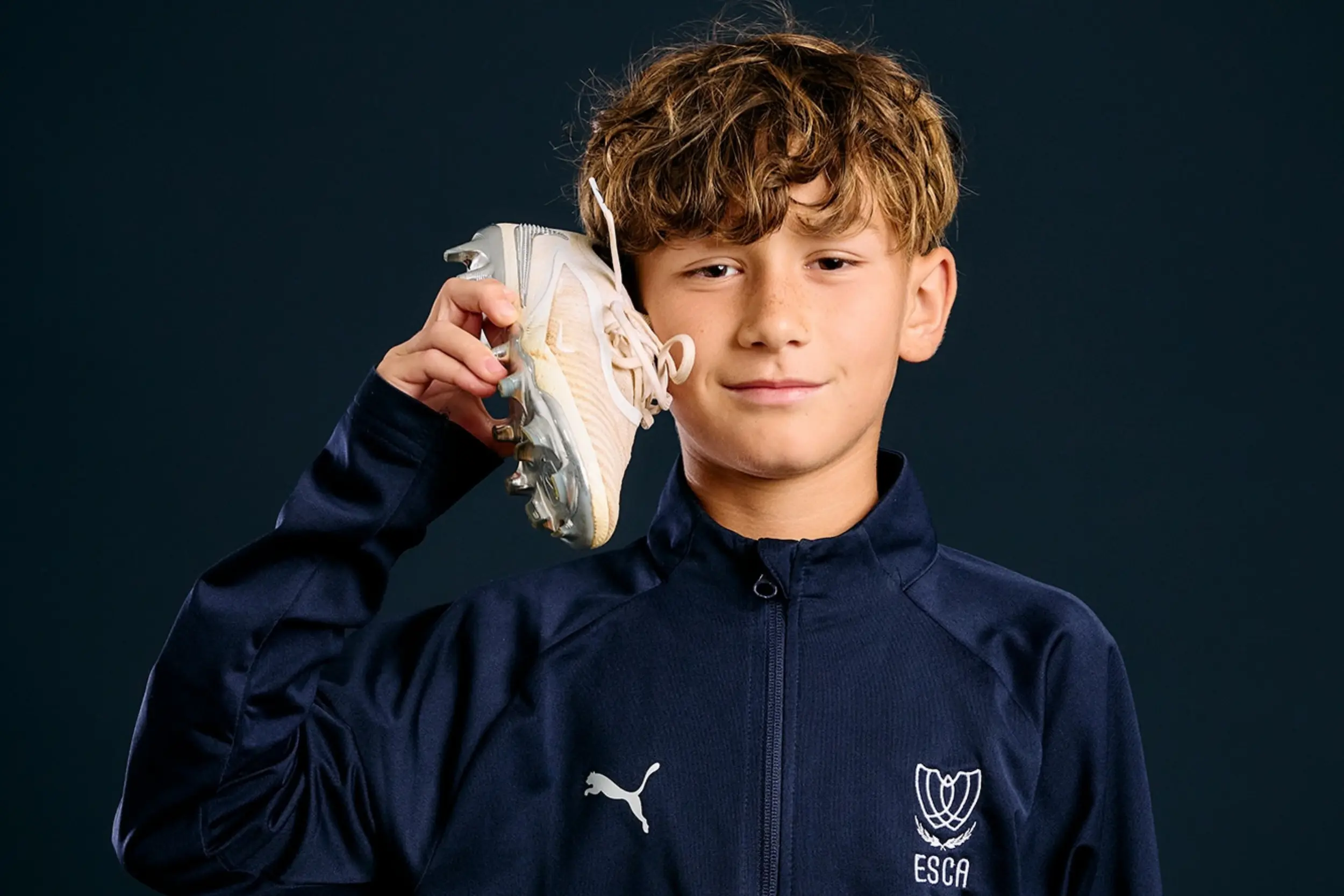 Smiling young woman with curly hair wearing a white ESCA Football Academy shirt against a dark background.