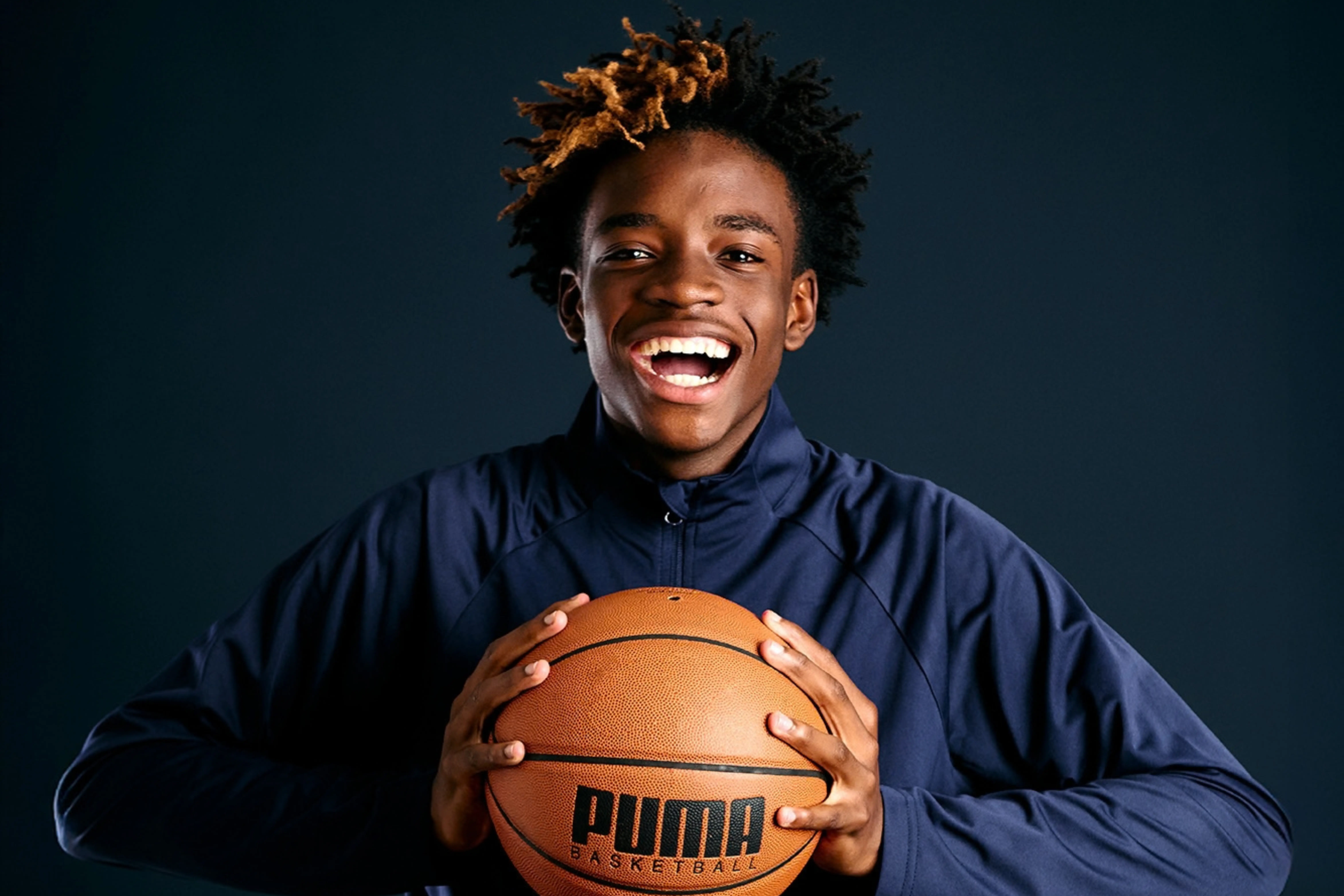Smiling young man with short, partially bleached hair holding a Puma basketball against a dark background, wearing a navy blue jacket.