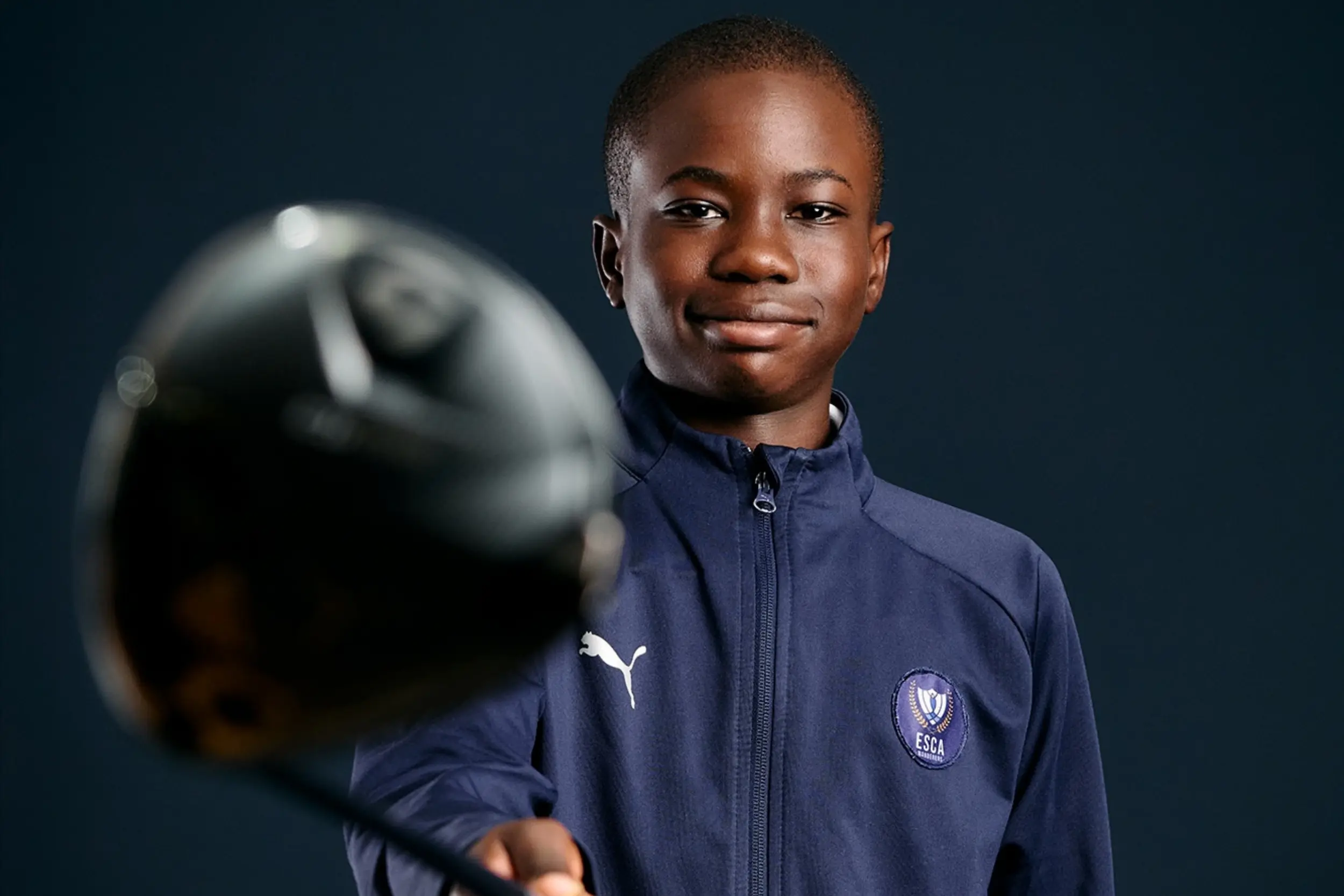 Smiling young boy wearing a navy blue Puma zip-up jacket with ESCA logo holding a golf club toward the camera against a dark background.