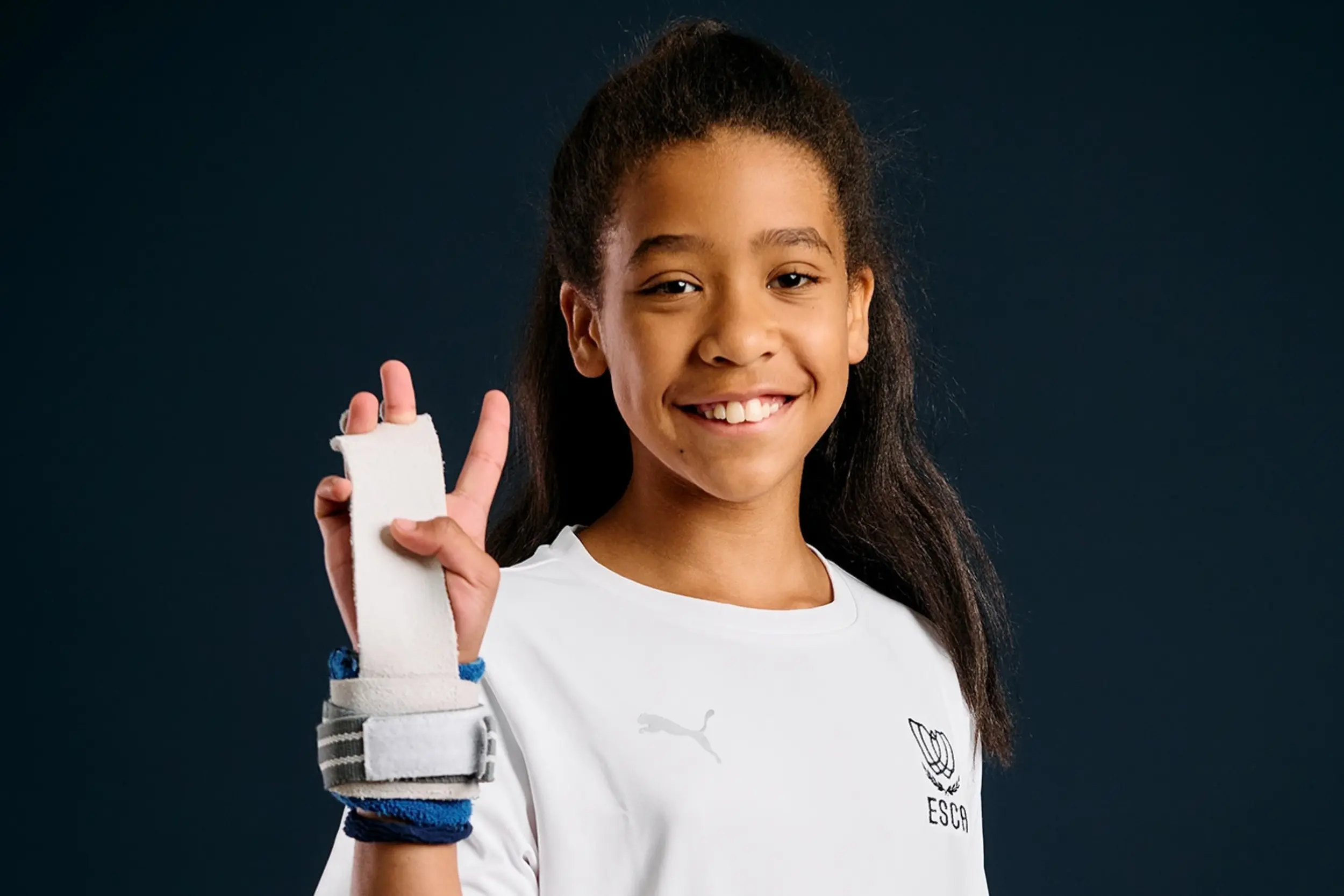 Smiling young girl wearing a white Puma shirt with ESCA logo, holding up her hand covered with a white wrist support against a dark background.