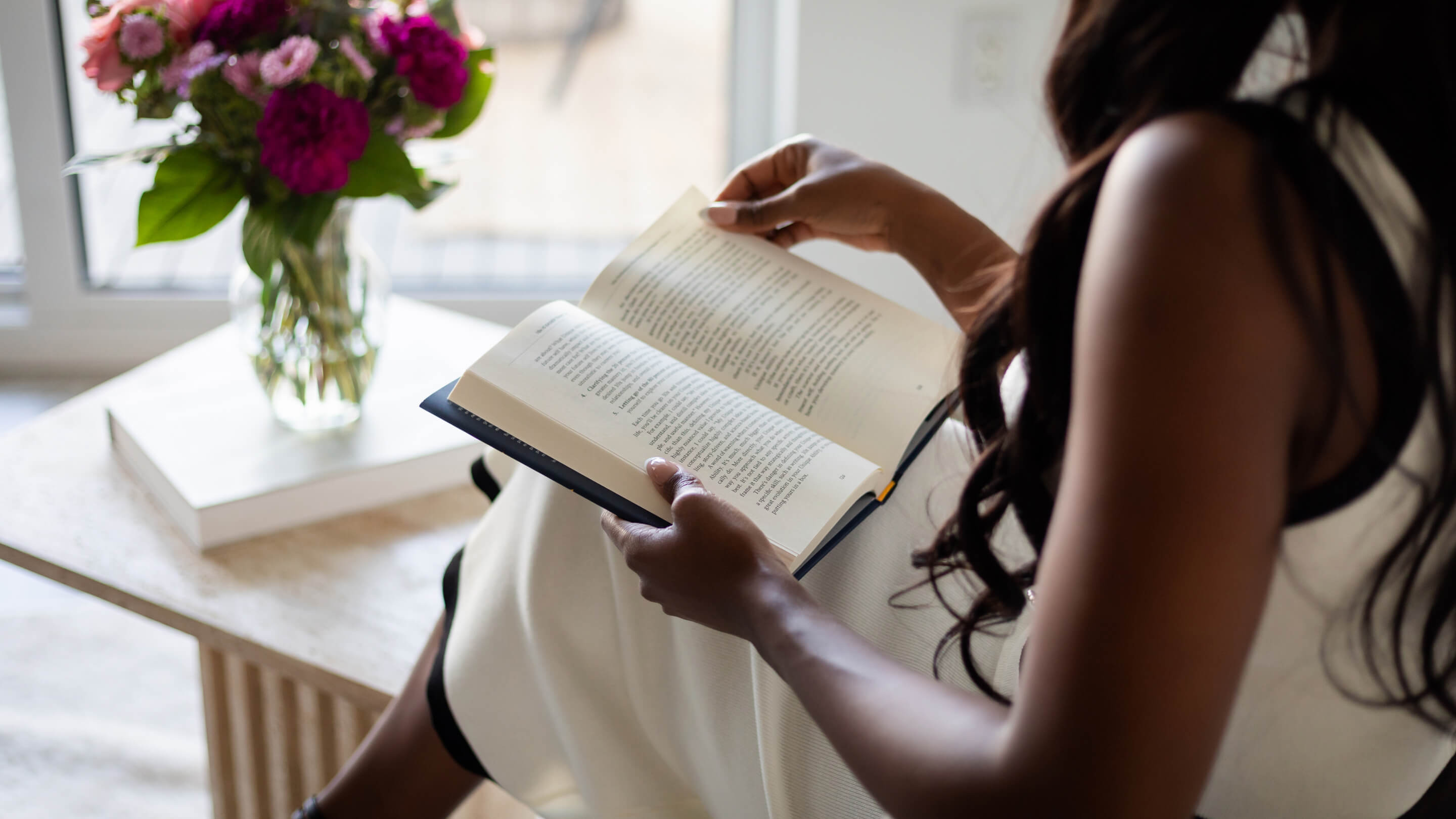 Person in white dress reading a book beside a table with a flower vase and a closed book.