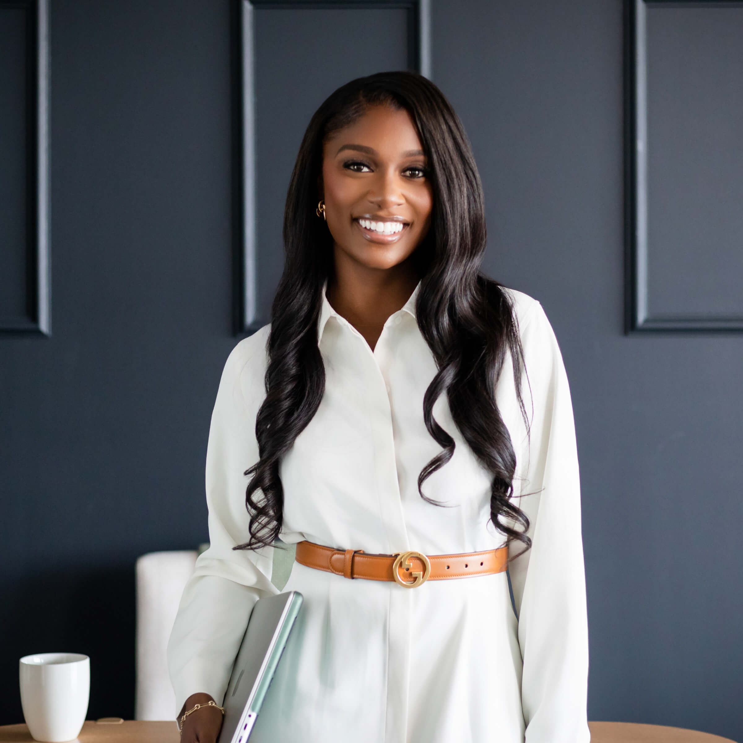 Smiling woman in white dress holding laptop, standing in modern office with dark paneled wall.