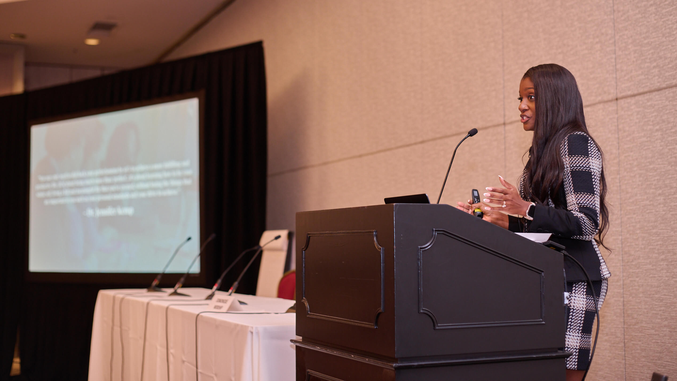 Woman in checkered suit speaking at a podium with a microphone in a conference room.