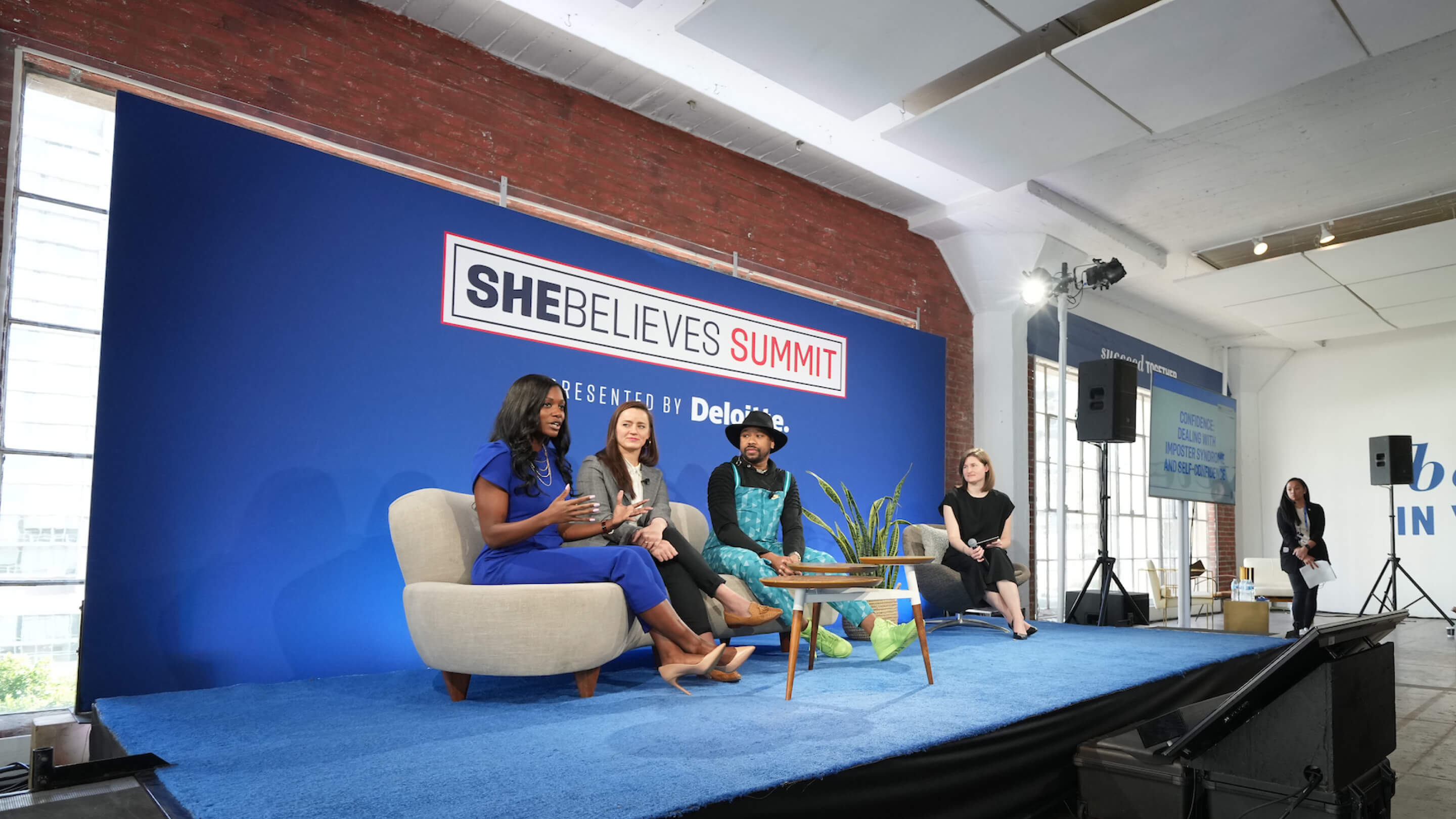Panel of four people seated on stage at She Believes Summit with blue backdrop and audience area.