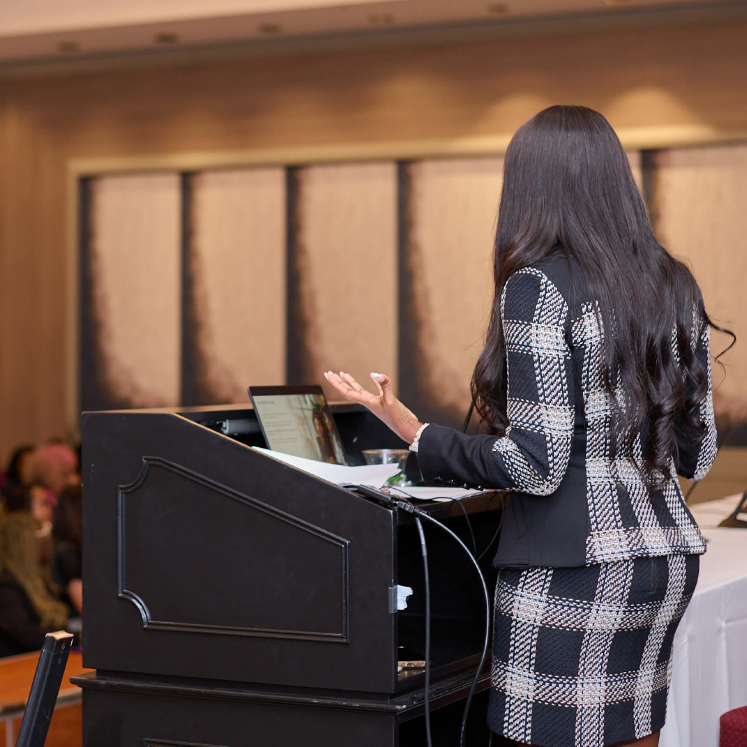 Woman in a plaid suit giving a presentation at a podium with a laptop and papers.