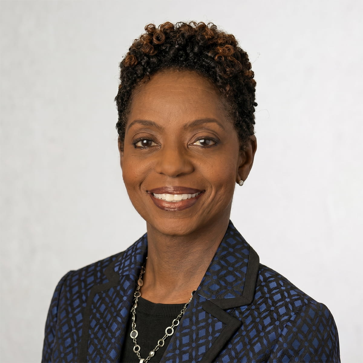 Woman with short curly hair in a patterned blue jacket smiling against a plain light background.