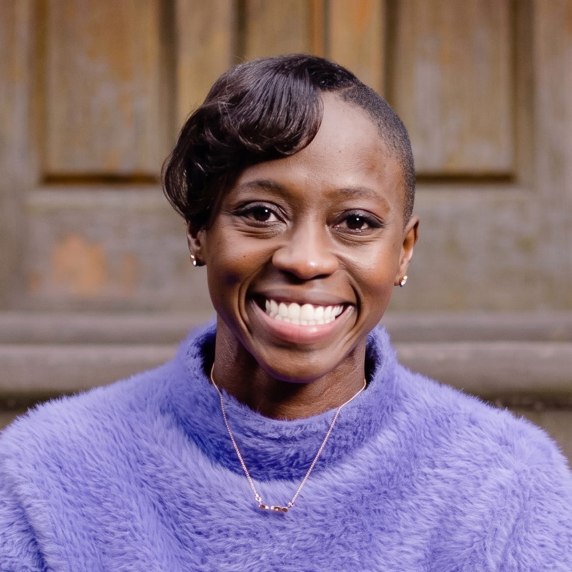 Smiling woman with short styled hair wearing a fuzzy purple sweater and a delicate necklace.