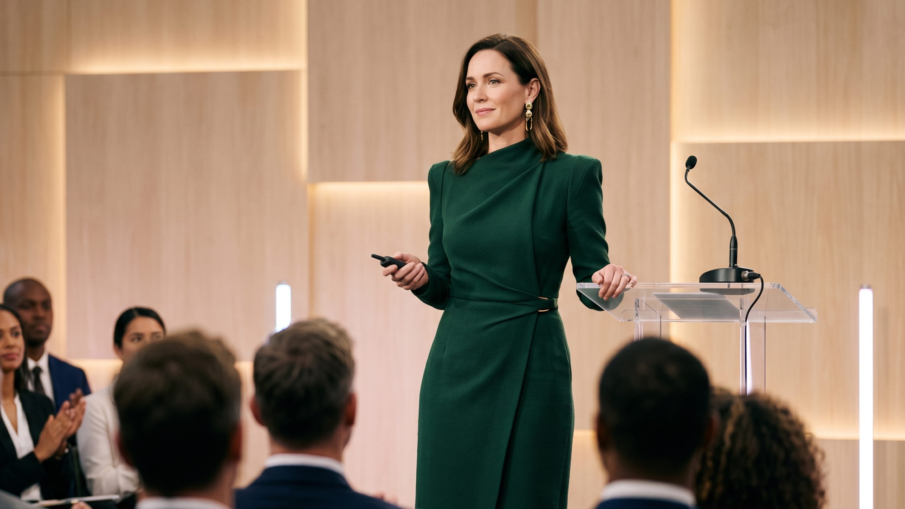 Woman in green dress speaking to an audience in a modern conference room with a clear podium.