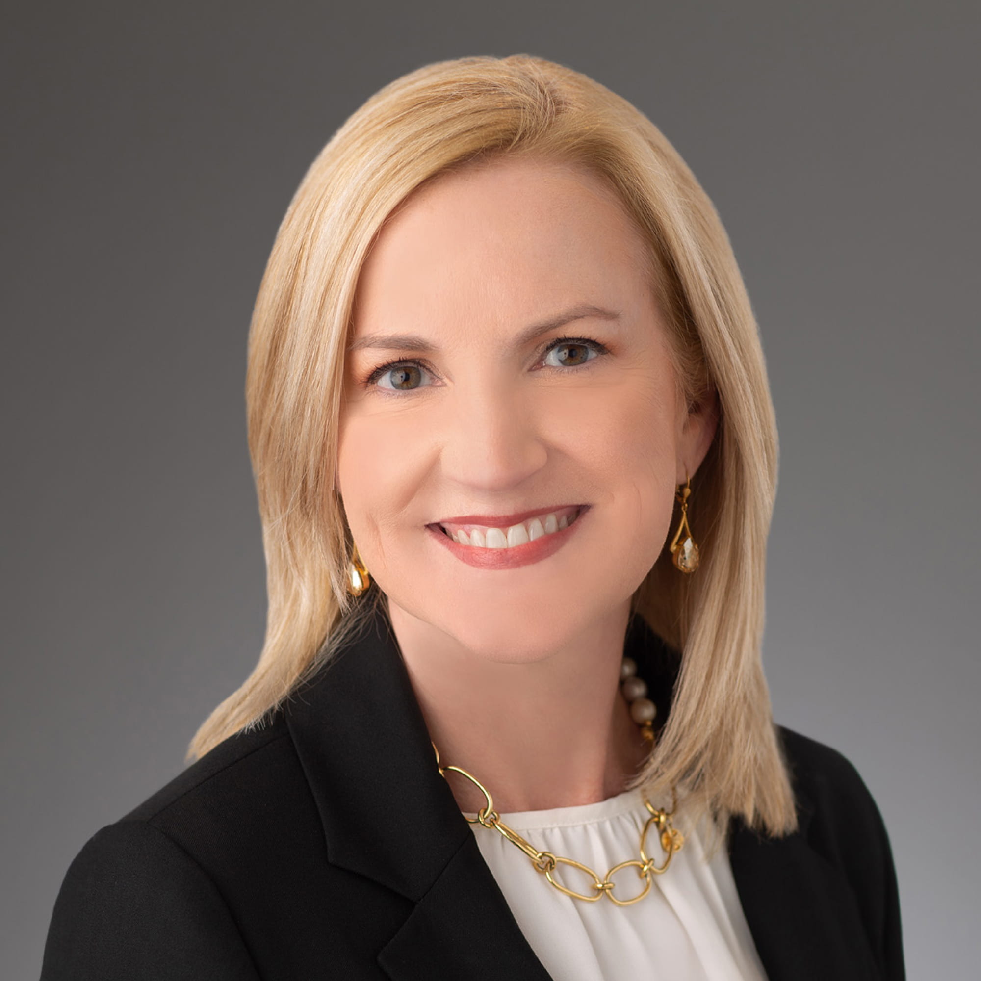 Smiling blonde woman in black blazer, white top, gold chain necklace, and drop earrings against gray background.