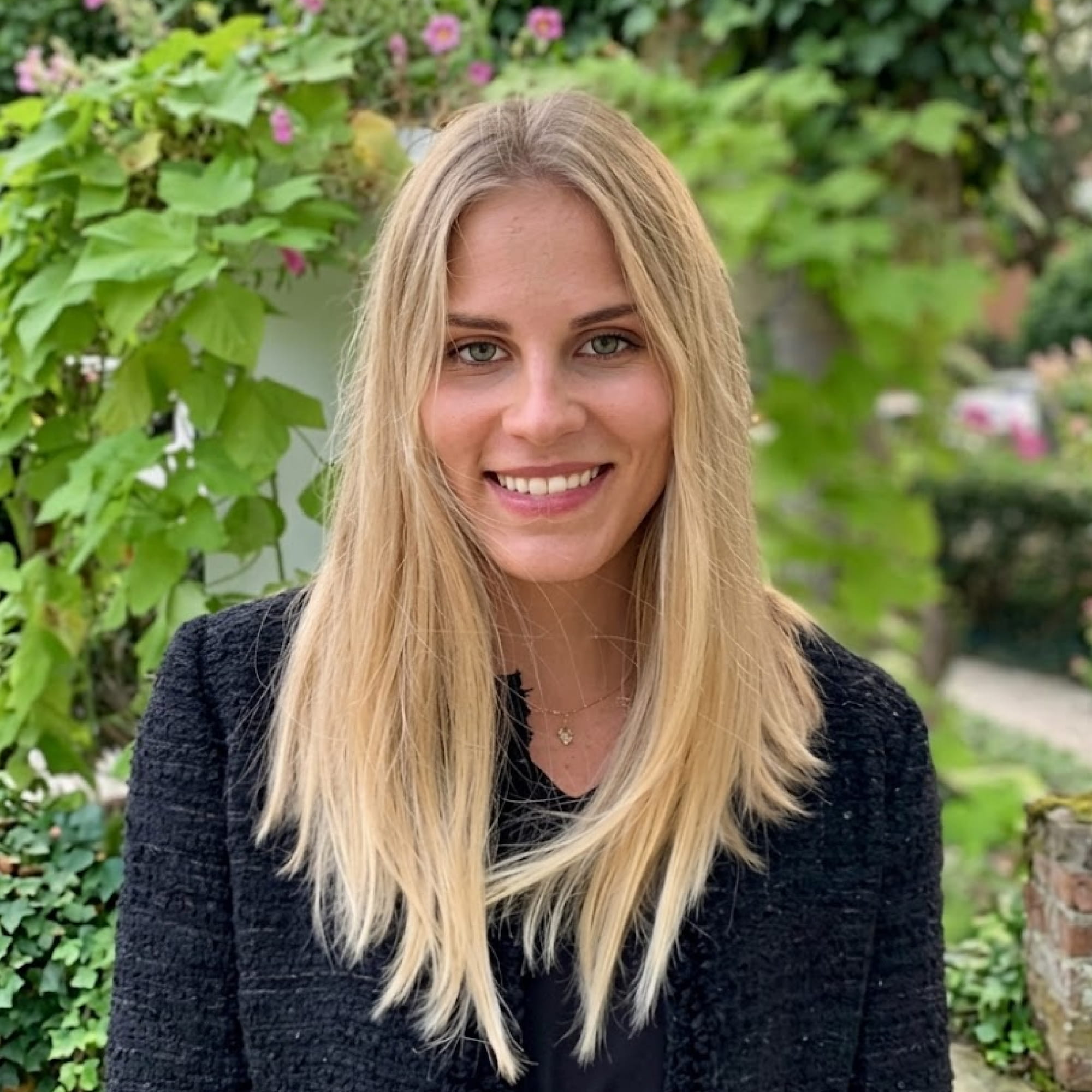Smiling blonde woman in black jacket with green plants and flowers in the background.