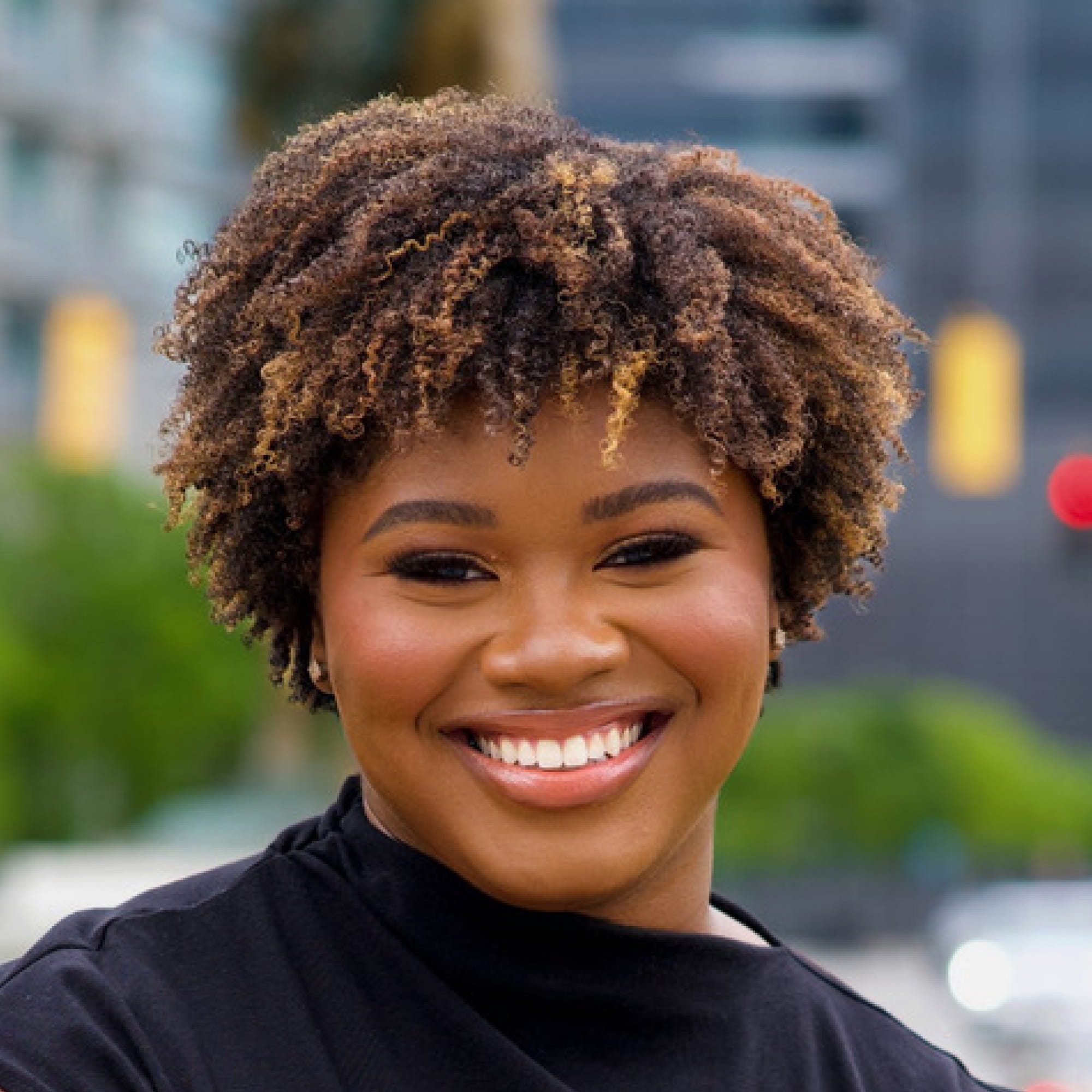 Smiling woman with curly hair wearing a black top in an urban outdoor setting.