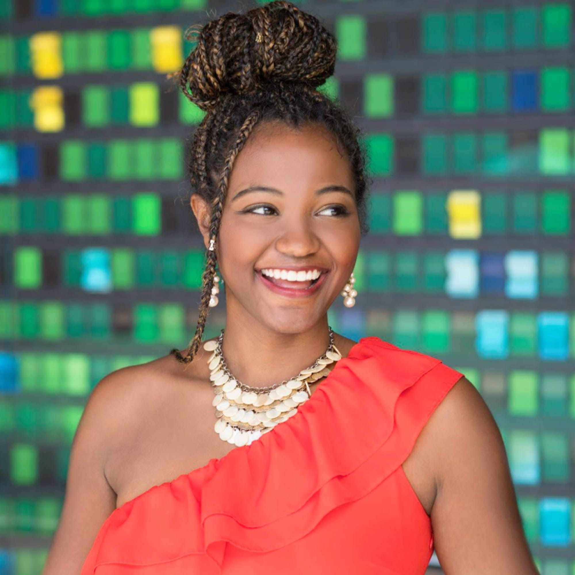 Smiling woman with braided updo wearing red off-shoulder top and layered necklace against green mosaic wall.