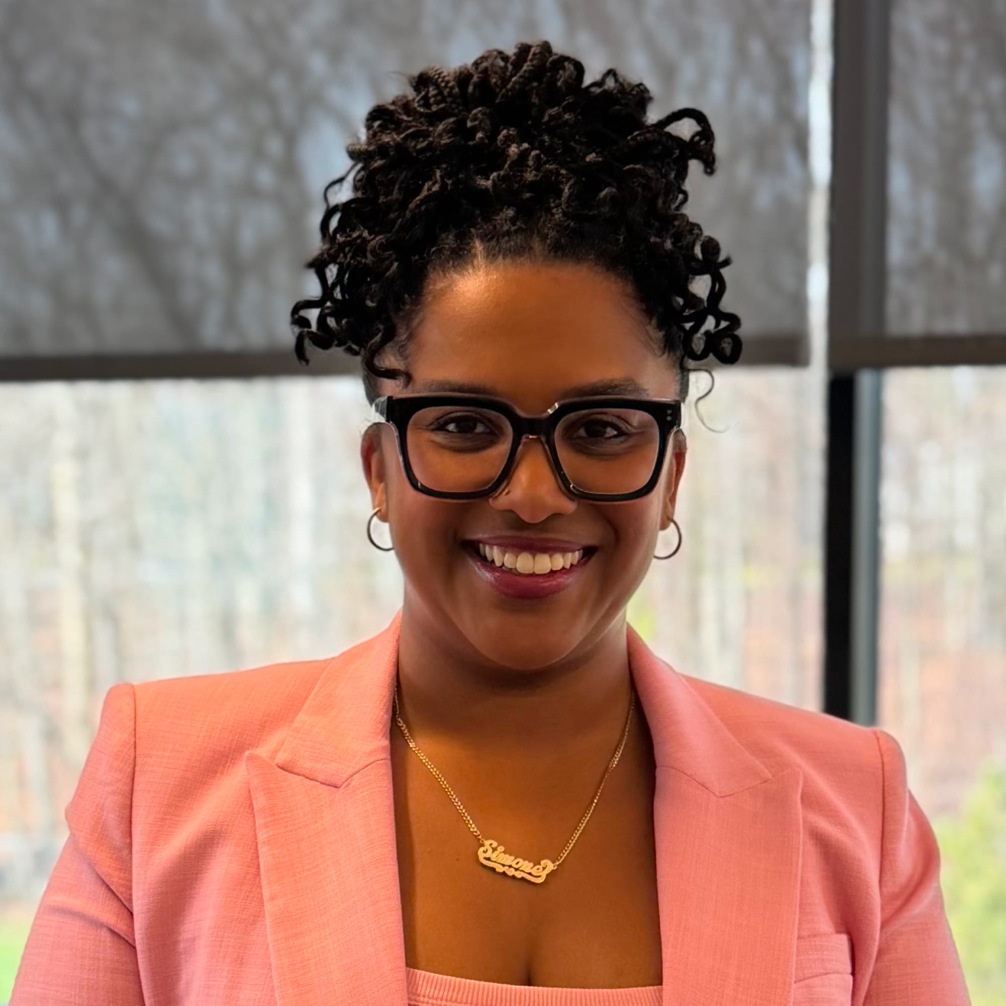 Smiling woman with curly updo, black glasses, pink blazer, and gold name necklace in front of window.