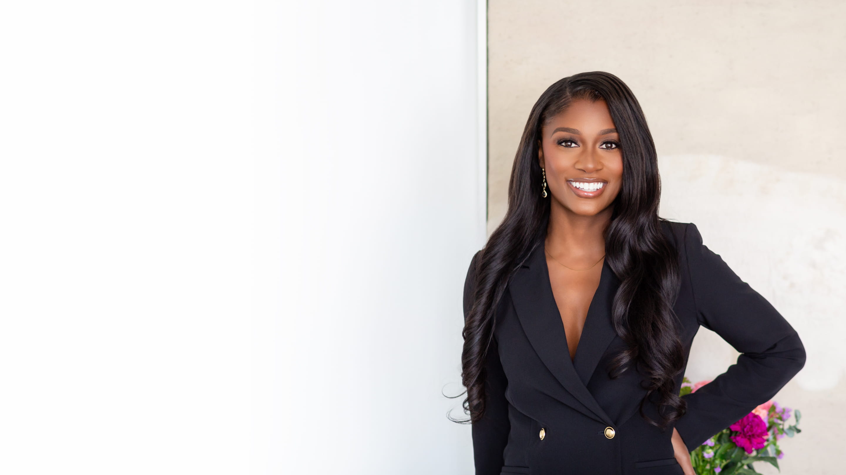 Smiling woman with long wavy hair wearing black blazer standing by a white wall with flowers nearby.