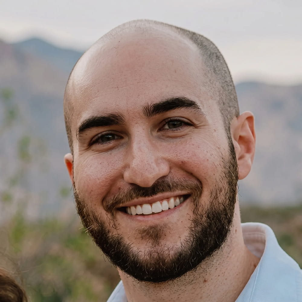 Smiling bald man with beard outdoors with blurred mountains in the background.