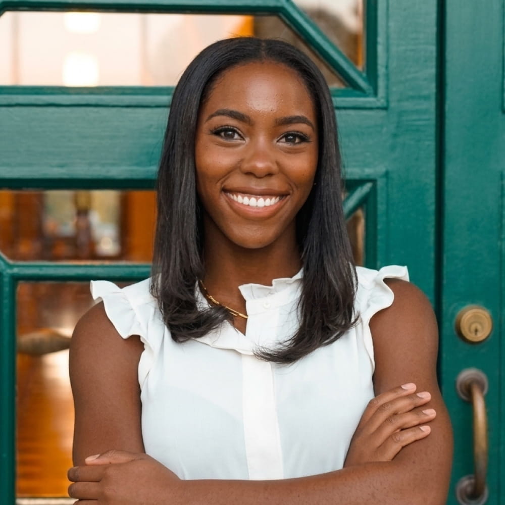 Smiling woman with crossed arms wearing a white sleeveless top standing in front of a green door.