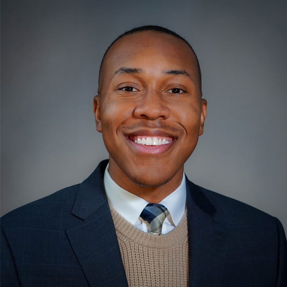 Smiling man wearing navy suit, brown sweater, white shirt, and patterned tie on gray background.