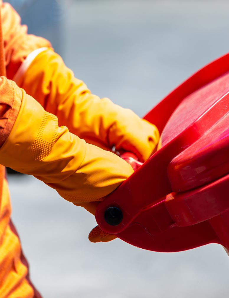 Person wearing orange gloves and clothing handling a red waste disposal bin lid.