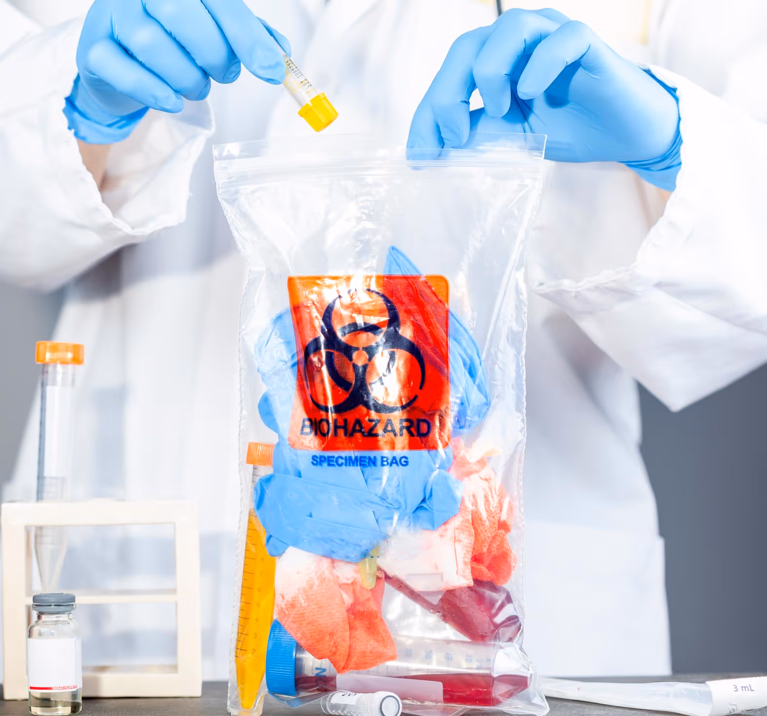 Gloved hands in lab coat placing a yellow-capped test tube into a transparent biohazard specimen bag containing gloves and medical waste.