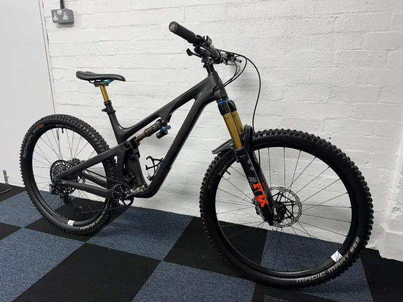 Black full-suspension mountain bike with Fox suspension forks against a white brick wall.