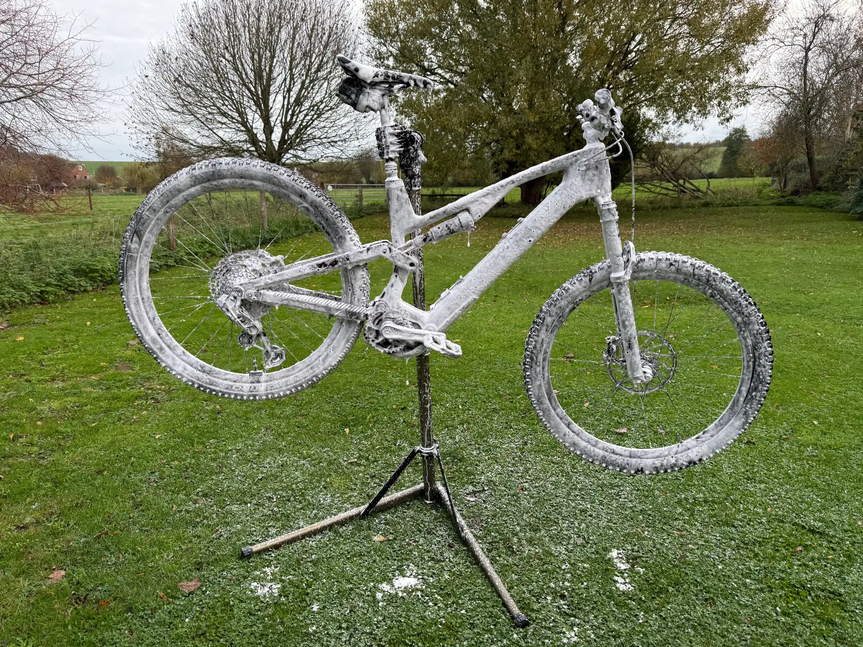 Mountain bike covered in foam, suspended on a repair stand outdoors on green grass with trees in the background.
