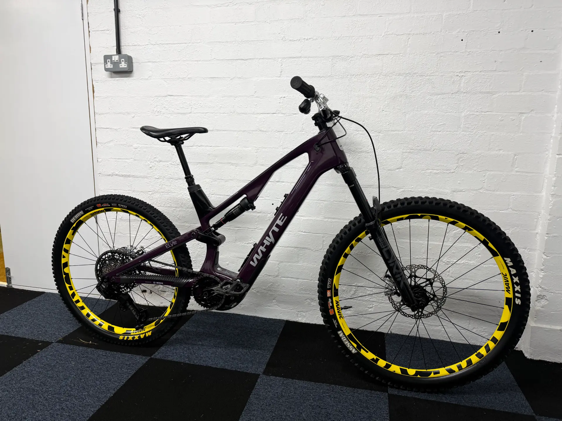 Purple Whyte mountain bike with yellow-rimmed tires leaning against a white brick wall on a checkered black and blue floor.