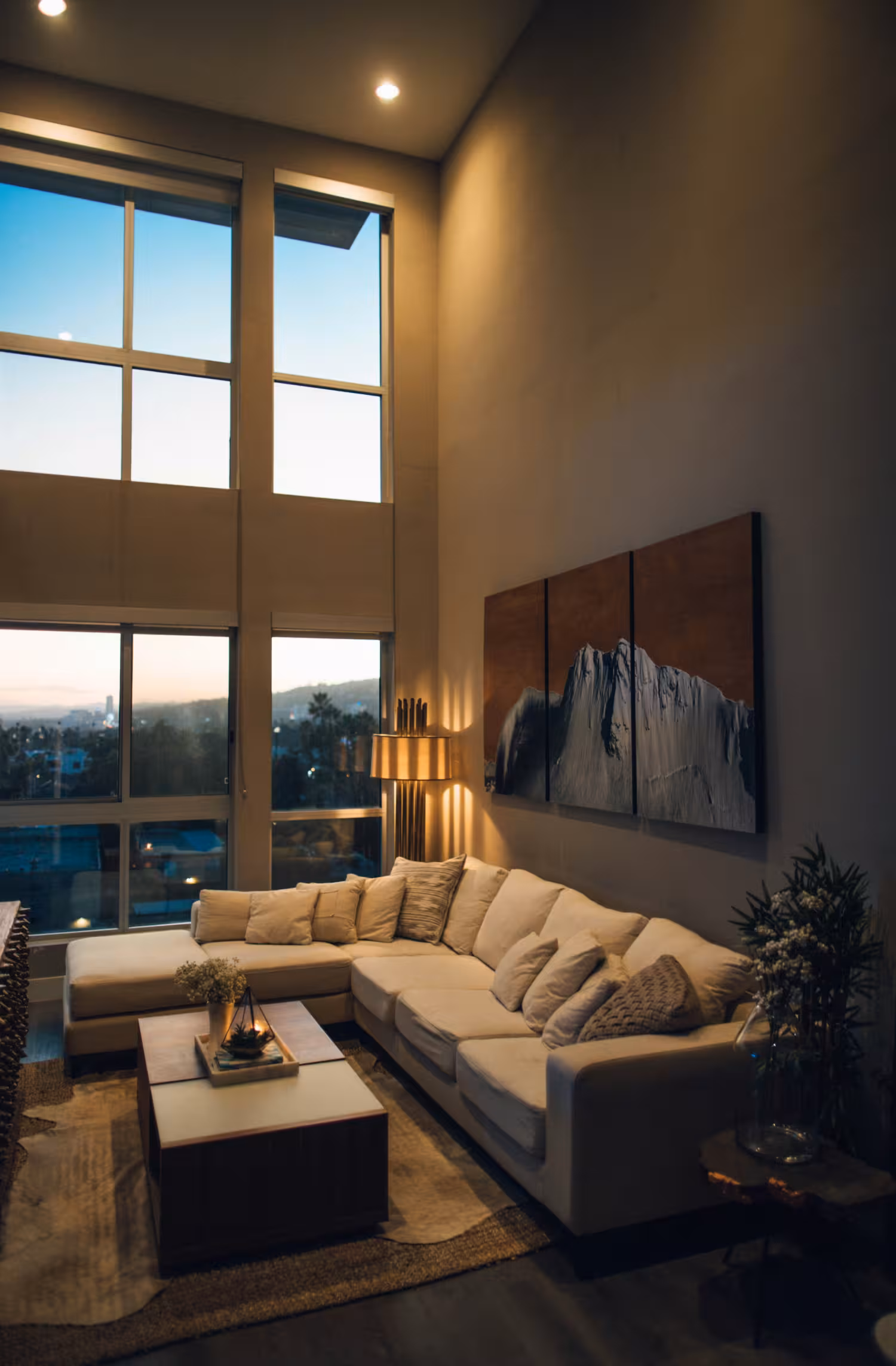 Sala moderna con sofá en forma de L, lámpara de pie y grandes ventanas al atardecer.