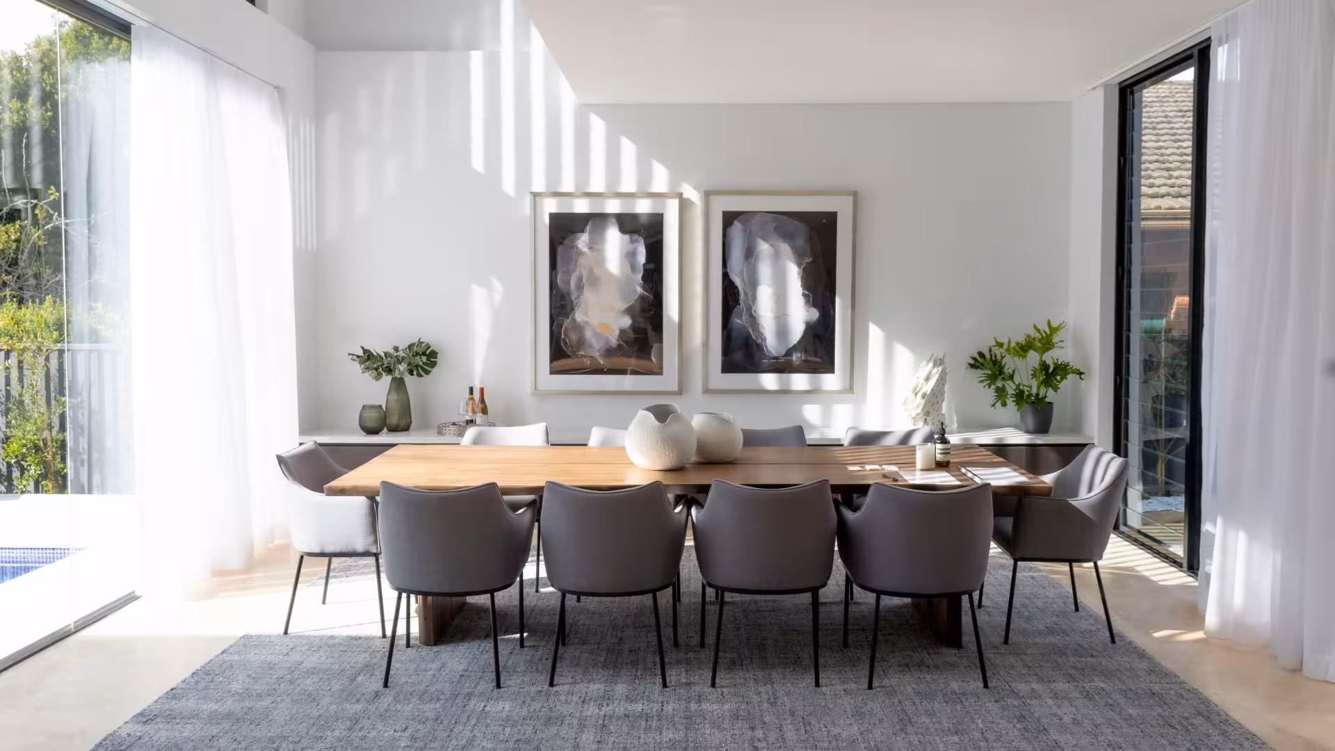 Comedor moderno con mesa de madera clara rodeada de ocho sillas grises, dos cuadros abstractos en la pared y grandes ventanas con cortinas blancas.