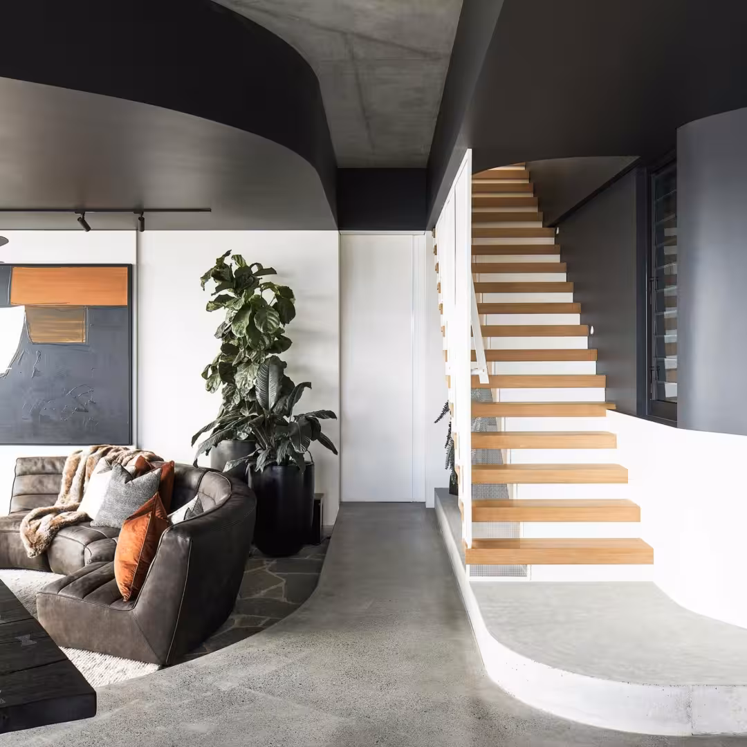 Sala de estar moderna con sofá marrón, plantas grandes y escaleras flotantes de madera con barandilla blanca.