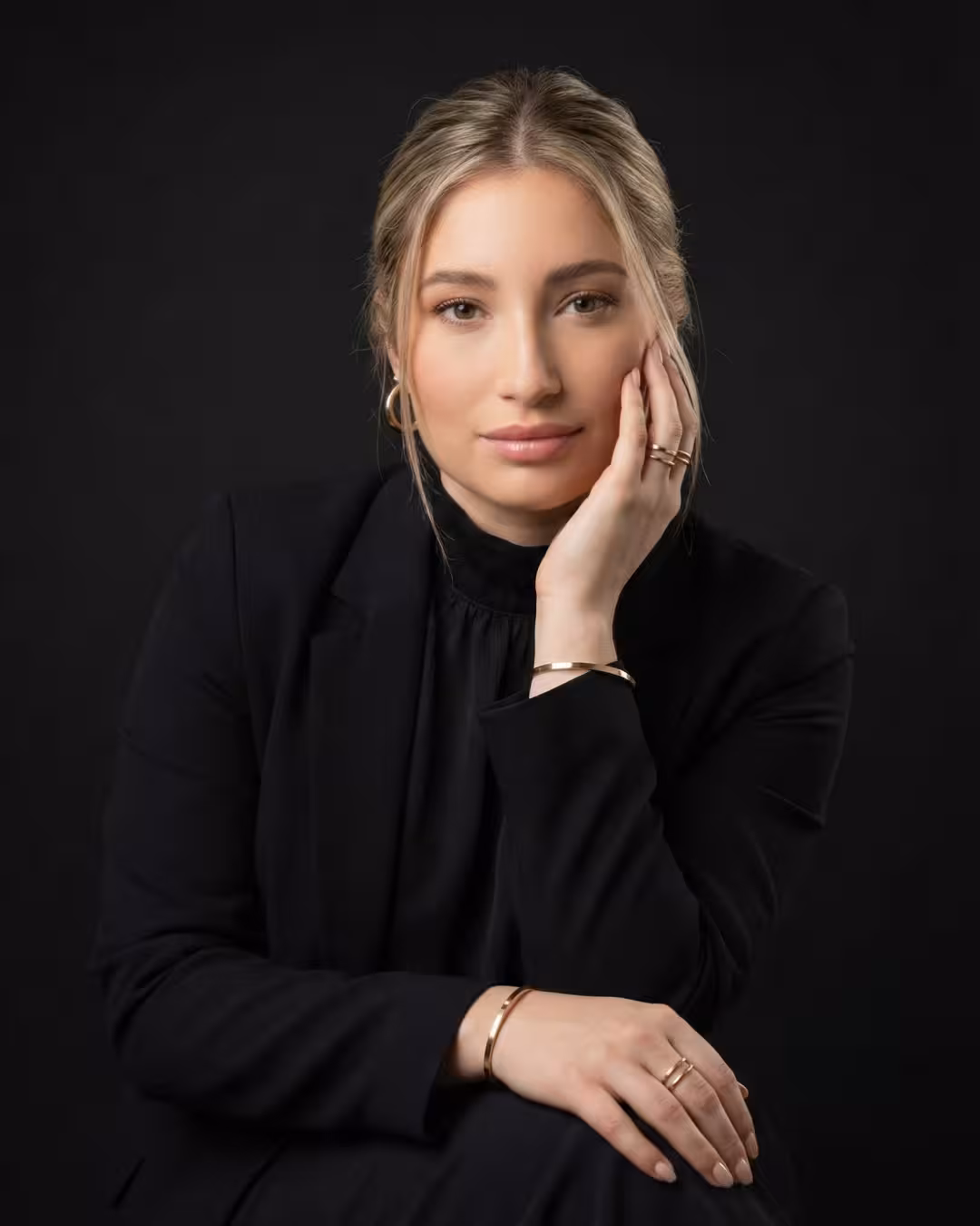 Mujer rubia con chaqueta negra y joyas doradas, posando pensativa con la mano en la cara sobre fondo negro.
