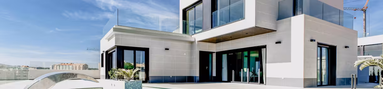 Casa moderna con grandes ventanales de vidrio y plantas alrededor bajo un cielo azul claro.