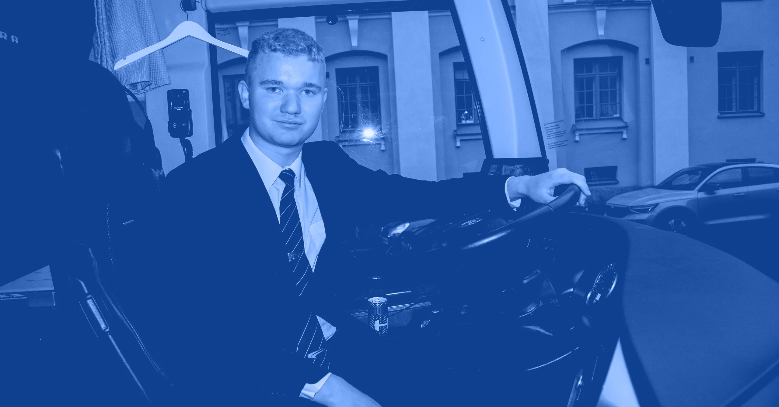 Young man in a suit sitting in the driver's seat of a vehicle, holding the steering wheel with a building visible through the window.
