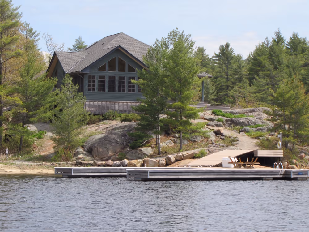 Lakeside cabin with large windows surrounded by pine trees and rocks, with a dock extending into the water and wooden chairs on it.