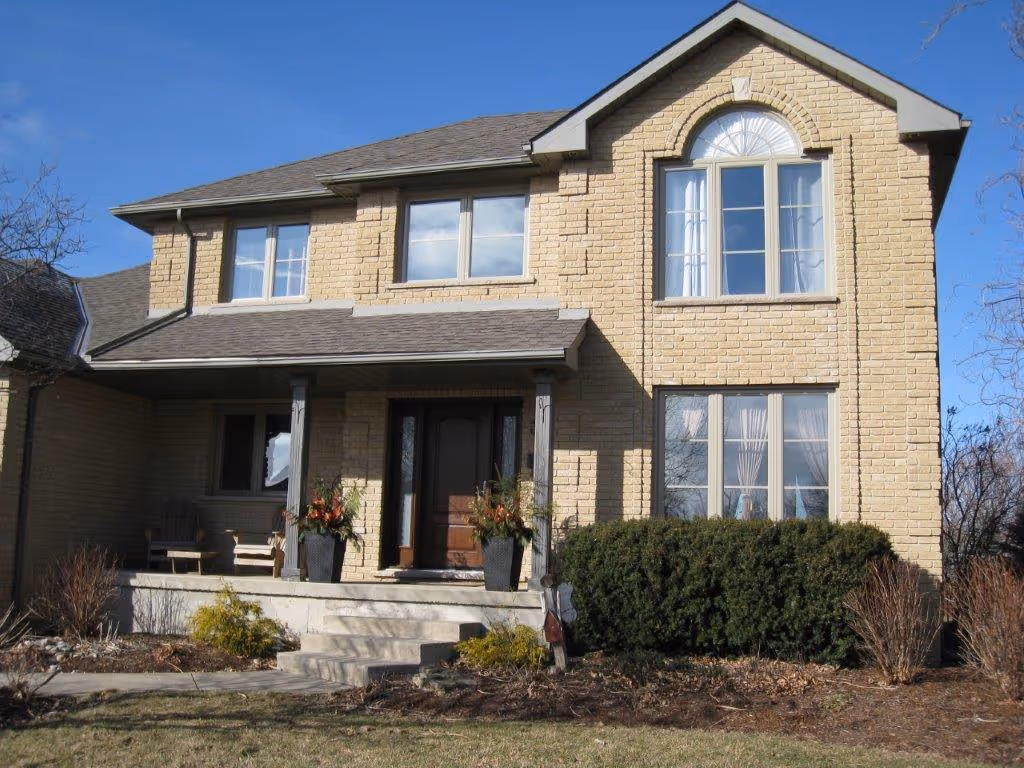 Two-story beige brick house with a covered front porch, wooden door, large windows, and shrubs in the front yard under a clear blue sky.