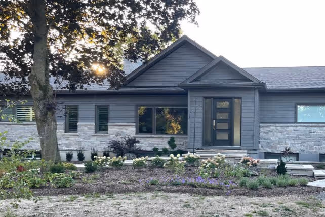 Modern single-story house with gray siding and stone accents, featuring a black front door and a large front garden with various plants and flowers.