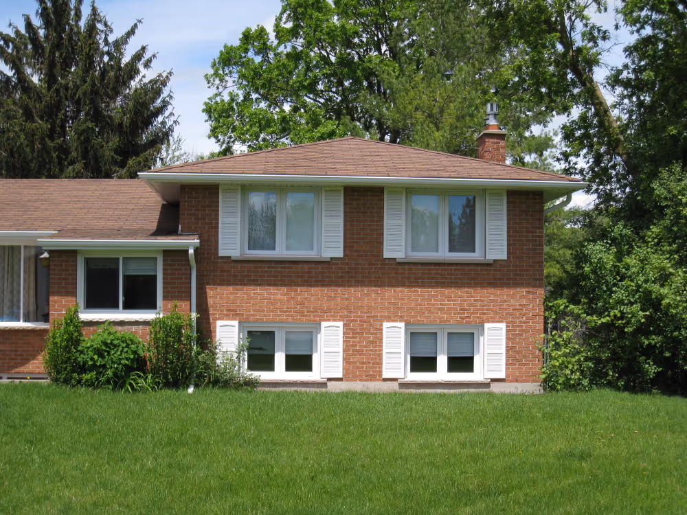 Two-story brick house with white shuttered windows and a brown shingled roof, surrounded by green grass and trees.