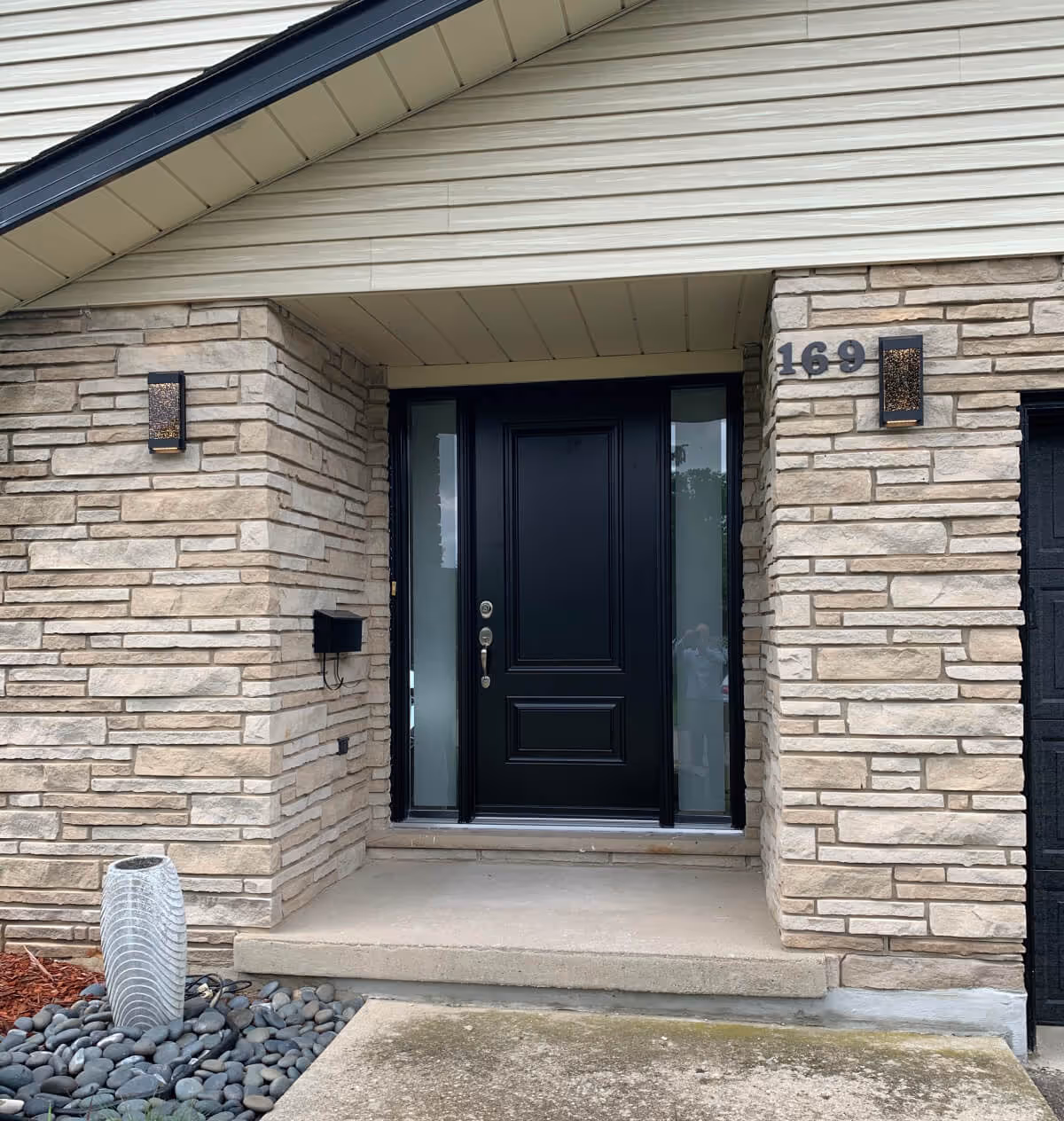 Front entrance with black door flanked by frosted glass panels, stone walls, and house number 169.