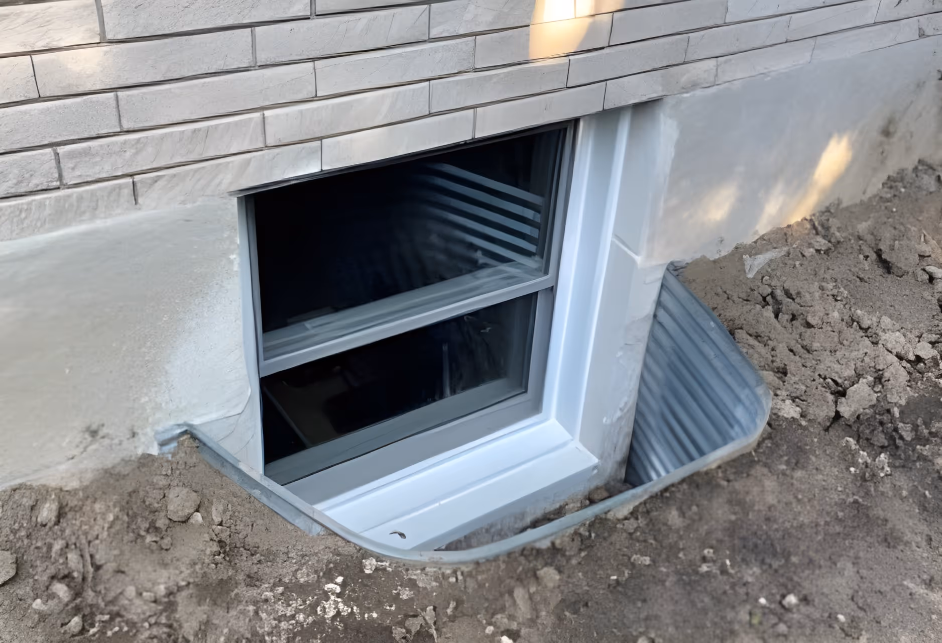Basement window with a newly installed white frame set in a wall surrounded by dirt and a corrugated metal window well.