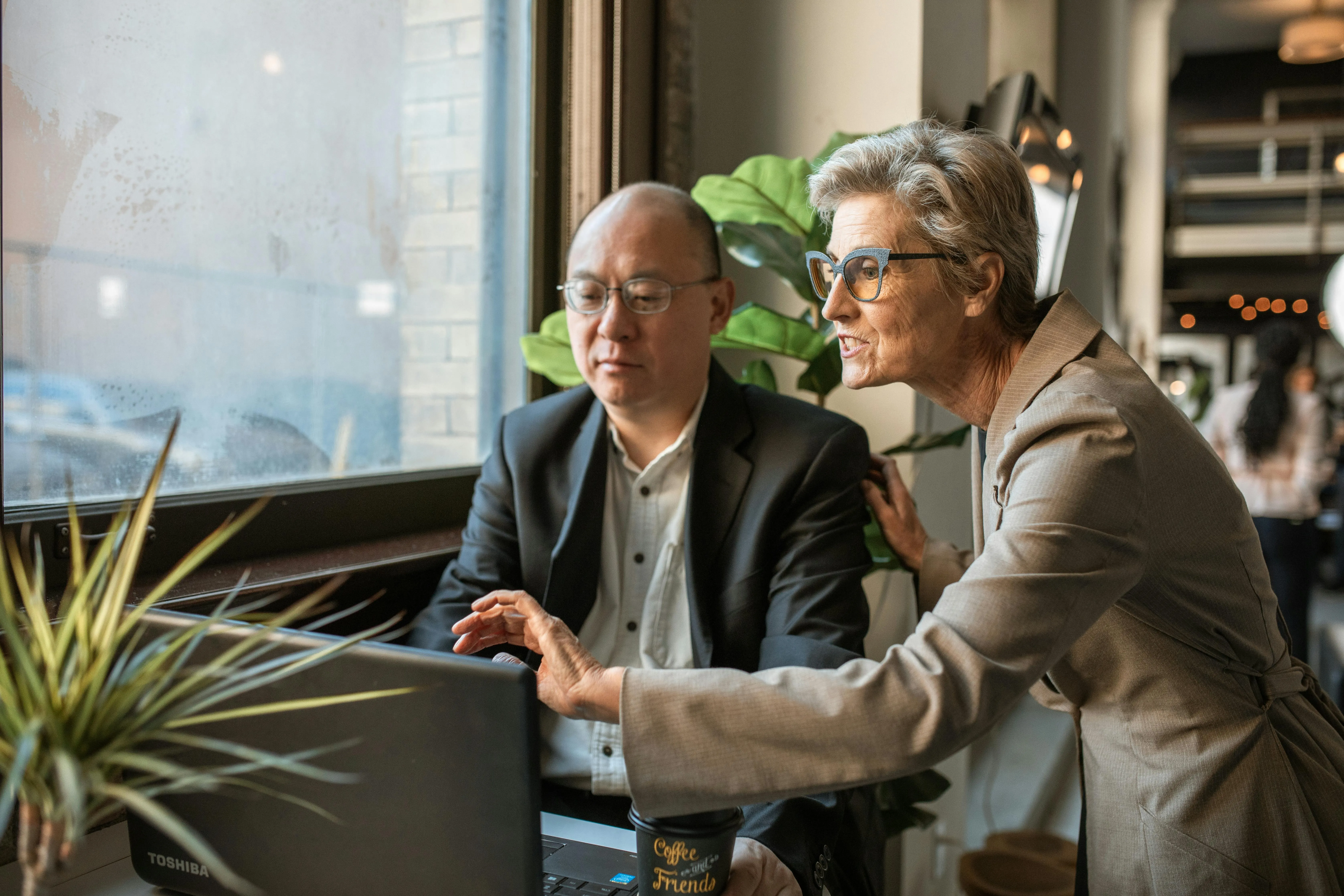 Two businesspeople collaborate over a Toshiba laptop near a window in a modern office setting.