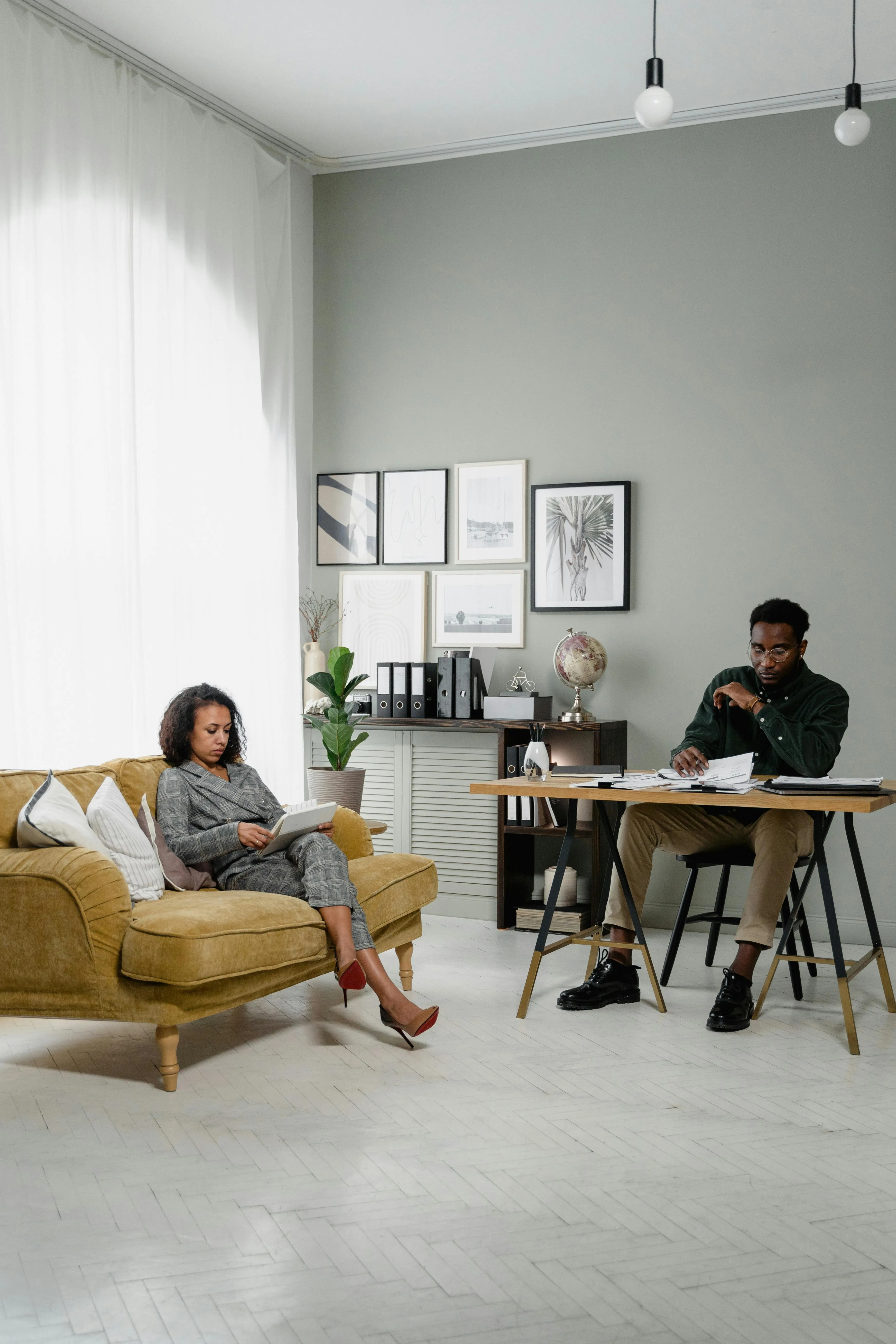 Woman in a gray suit reading a book on a mustard-colored sofa, man in a dark shirt sitting at a wooden desk reviewing papers in a modern, bright office.