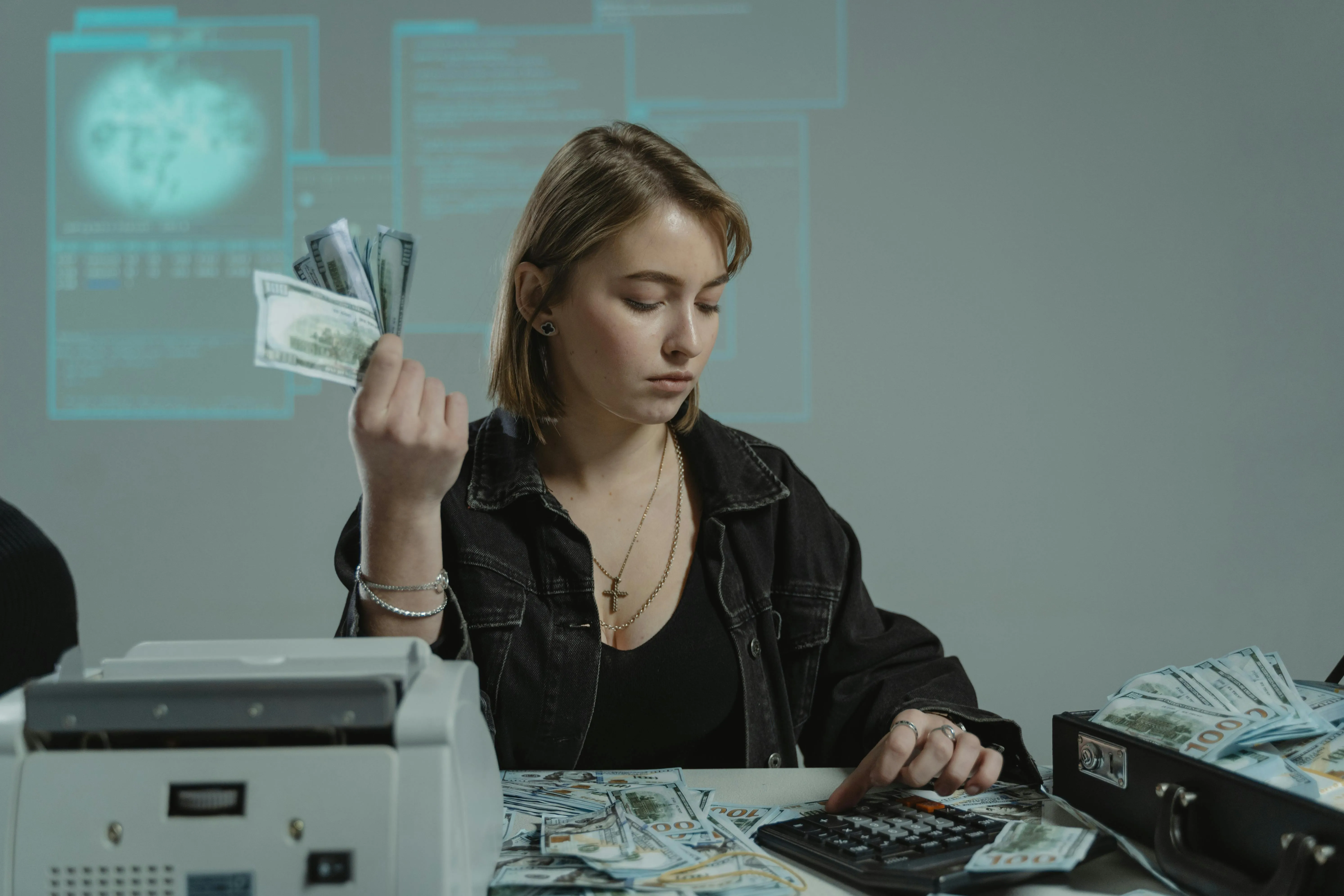 Young woman in black jacket holding US dollar bills, using a calculator, with cash and a briefcase on the table.