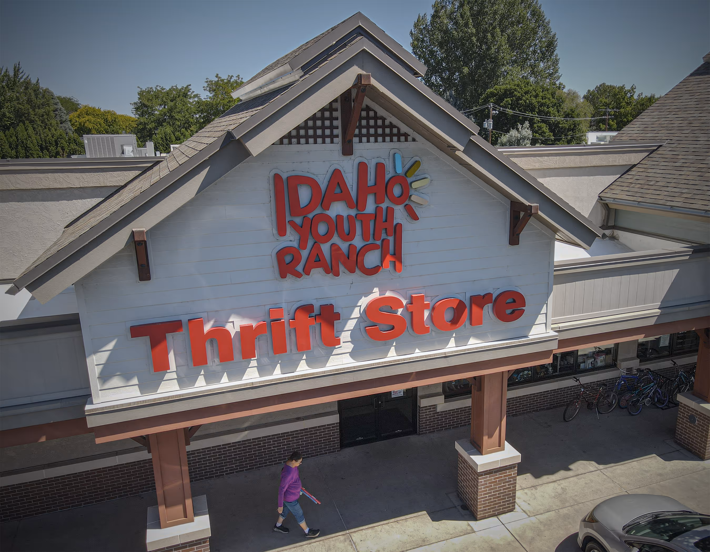 Exterior view of Idaho Youth Ranch Thrift Store with red signage; a person in a purple top and shorts walks on the sidewalk below.