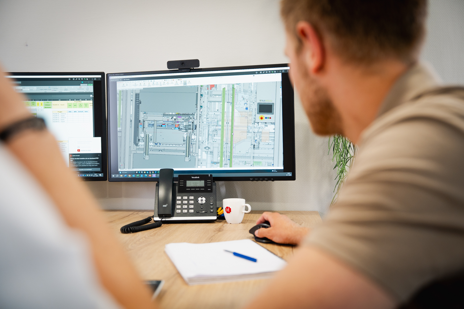 Man werkt achter een bureau met twee computerschermen waar technische tekeningen en spreadsheets zichtbaar zijn.