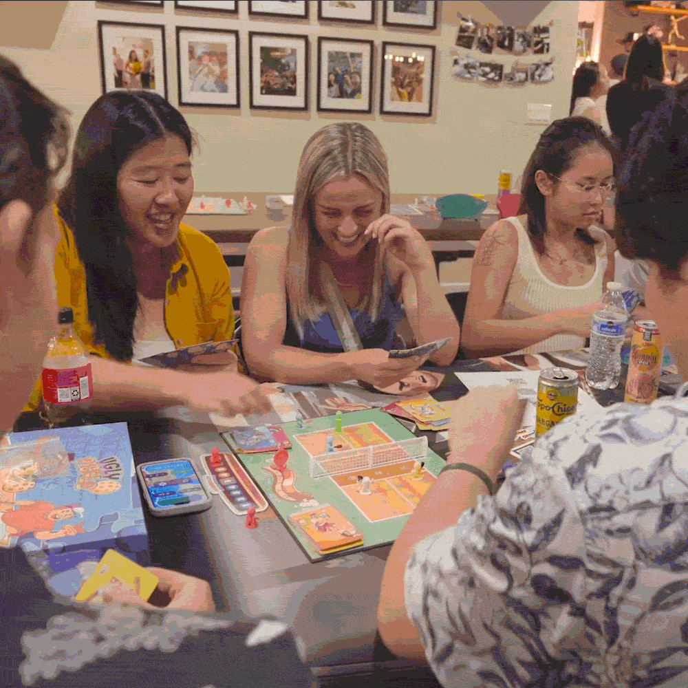Four people gathered around a table playing a colorful board game with cards and character pieces, smiling and interacting.