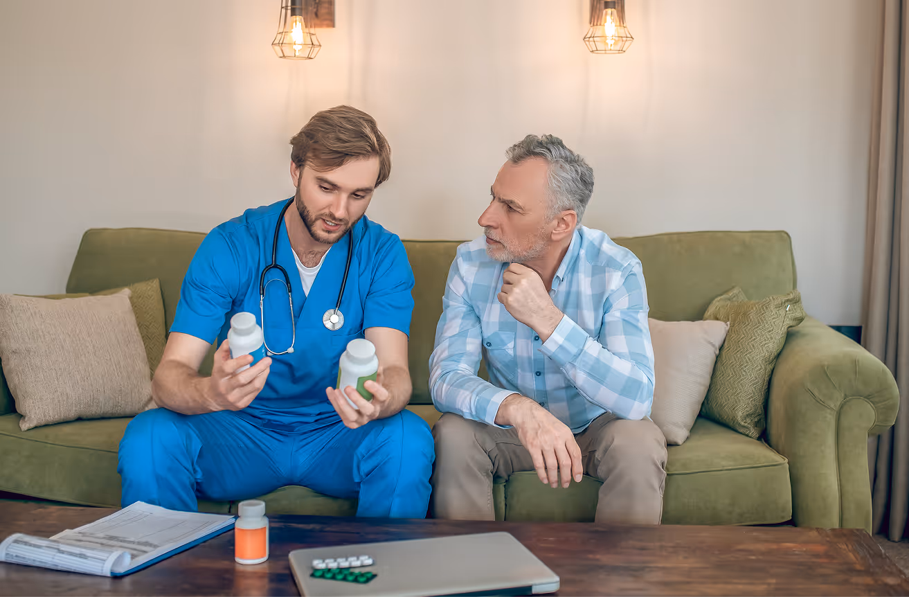 Male nurse in blue scrubs explaining medication bottles to an elderly man seated on a green sofa.